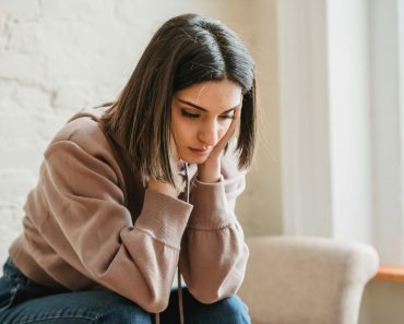 Unhappy female in casual wear touching face and looking down while sitting on sofa in light living room at home