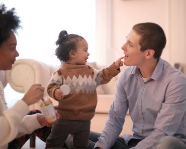 Side view of diverse parents in casual clothing sitting on floor and having fun with smiling toddler while gathering in cozy living room and having rest together