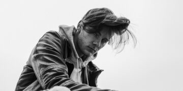 grayscale photo of man swearing leather jacket sitting on floor near building