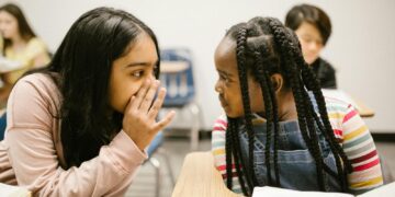 Two young girls whispering in a classroom setting, sharing a secret.