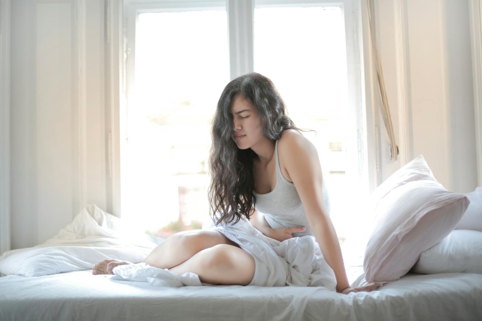 Woman sitting on bed holding her stomach showing signs of discomfort or pain indoors