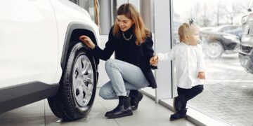 Full length of cheerful smiling mother in jeans holding hands with cute little girl and looking at car wheel in car showroom in daylight