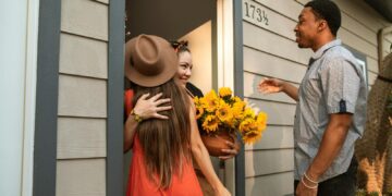Three friends greeting and hugging at a house entrance with sunflowers.