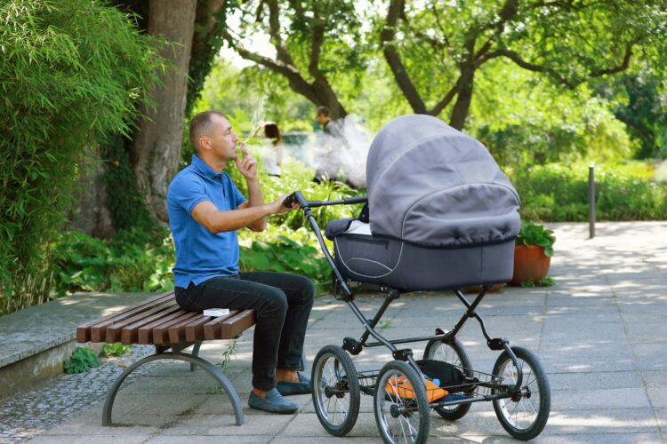 A man smoking a cigarette while sitting on a bench next to a stroller