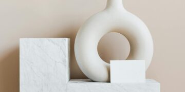 Composition of creative white ceramic vase in ring shape with empty postcard placed on white marble shelf against beige wall as home decoration elements or art objects