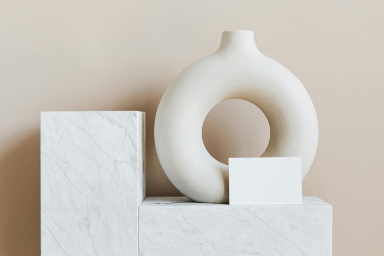 Composition of creative white ceramic vase in ring shape with empty postcard placed on white marble shelf against beige wall as home decoration elements or art objects