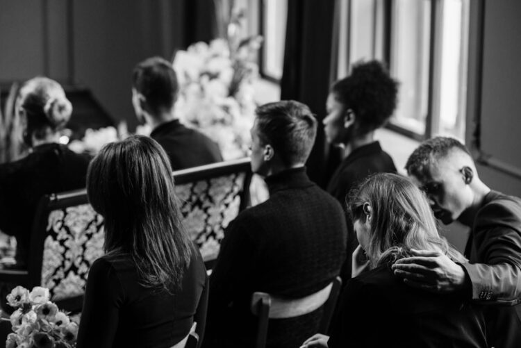 A somber grayscale image of mourners sitting indoors during a funeral conveying loss and sympathy