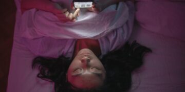A woman lying in bed at night browsing her smartphone under soft lighting.