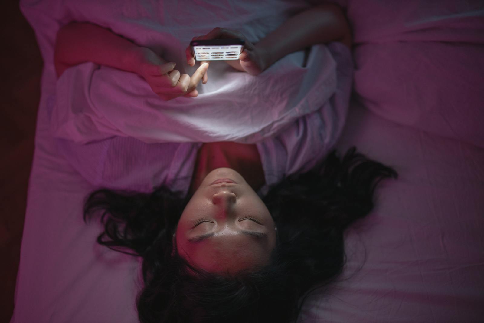 A woman lying in bed at night browsing her smartphone under soft lighting