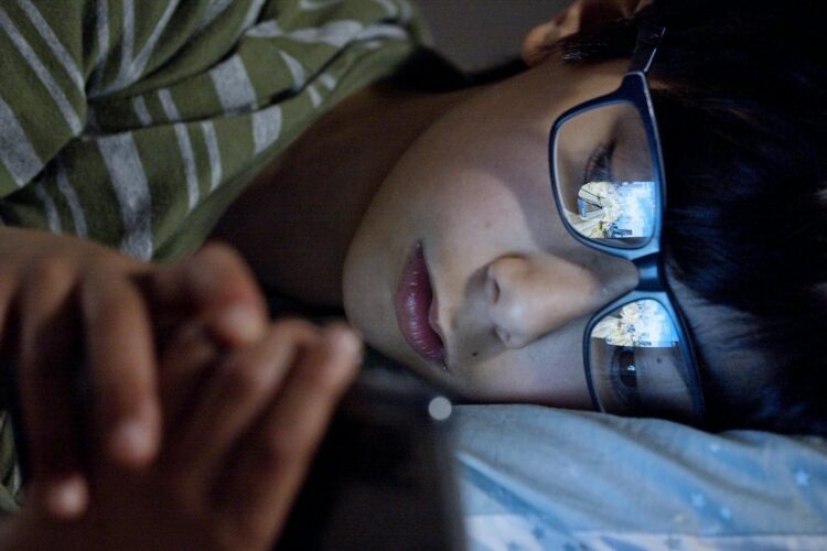 A child lying in bed at night using a smartphone with screen reflection on eyeglasses