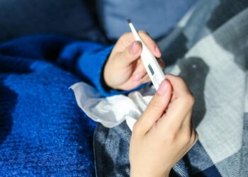 A woman checks a digital thermometer while lying indoors showing signs of illness