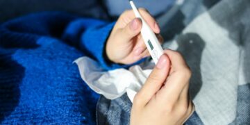 A woman checks a digital thermometer while lying indoors, showing signs of illness.