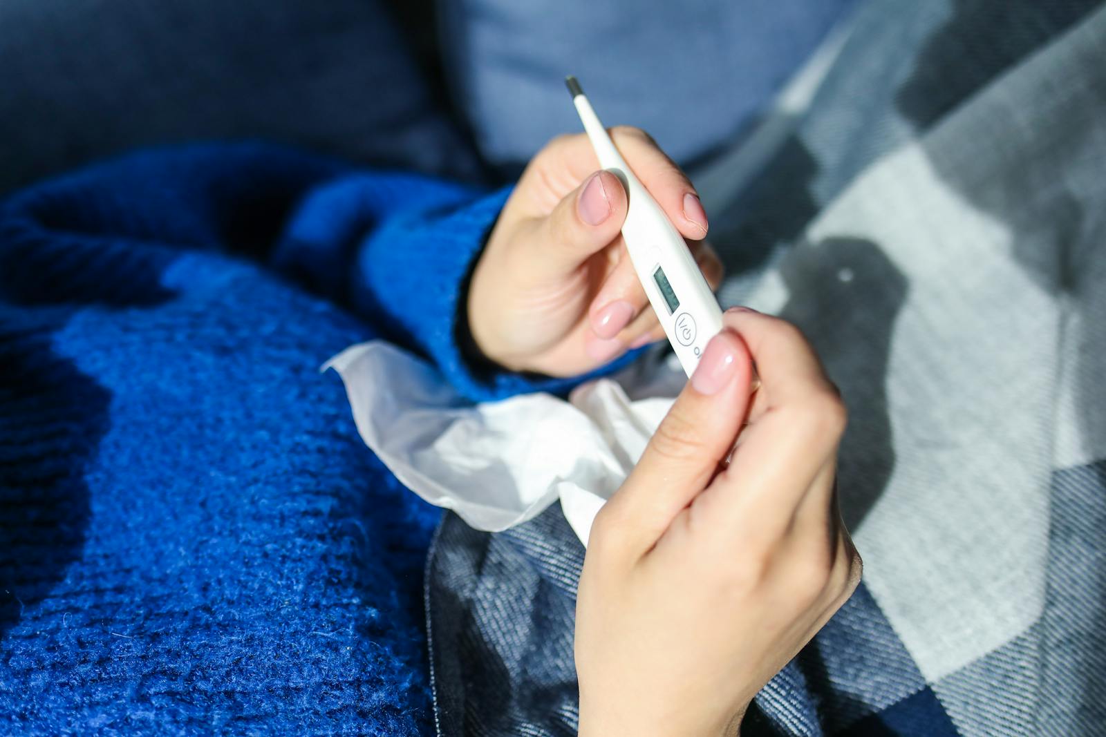 A woman checks a digital thermometer while lying indoors showing signs of illness
