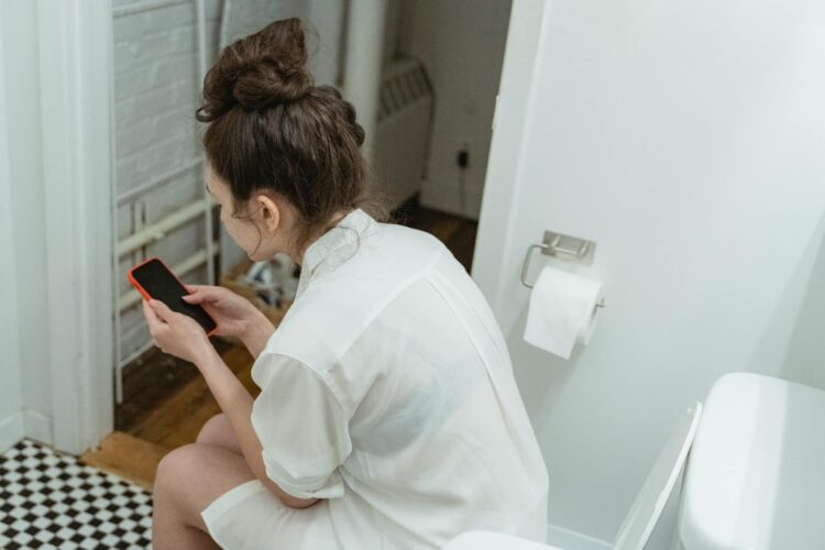 A woman wearing a white shirt uses a smartphone while sitting in a modern bathroom suggesting relaxation and comfort