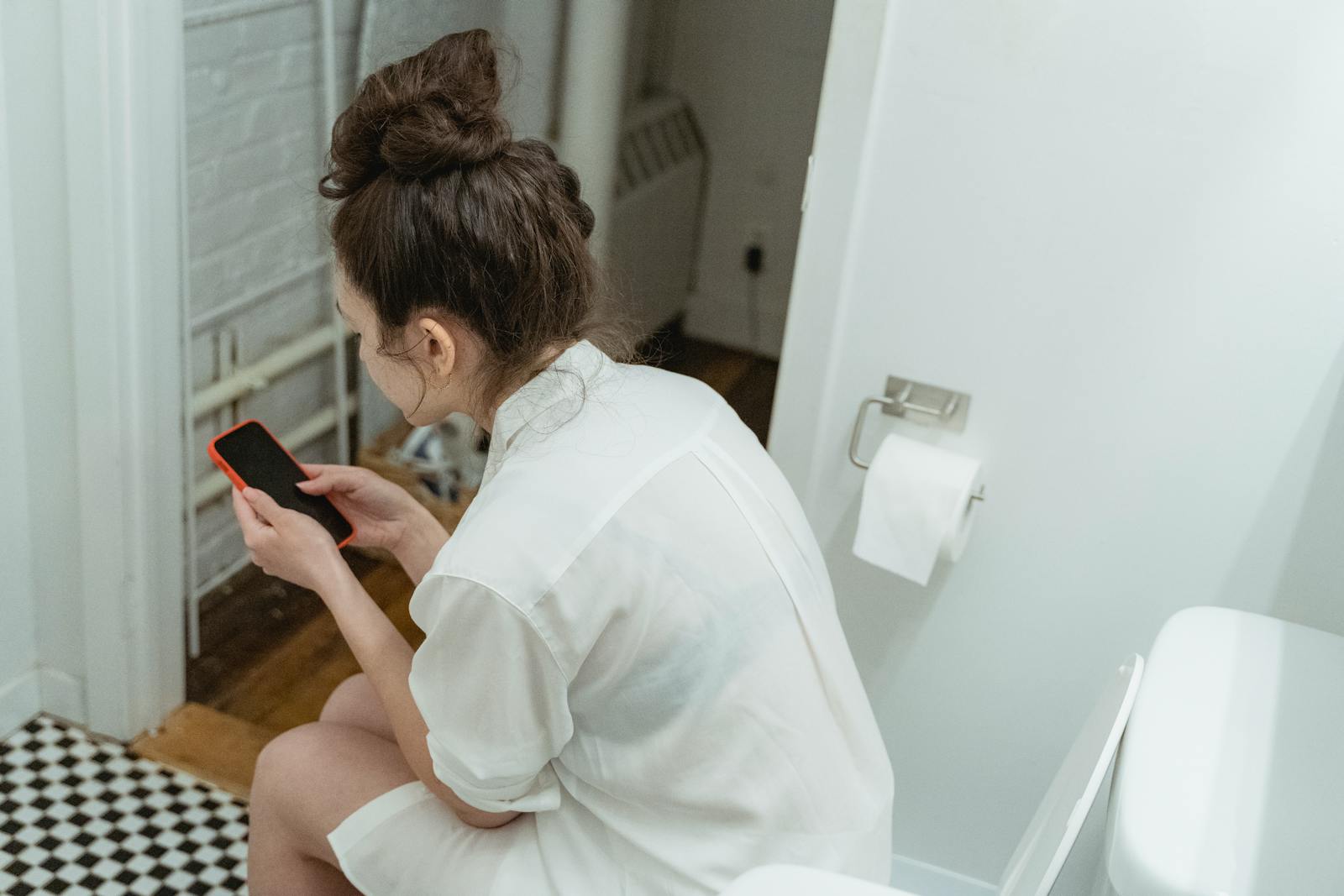 A woman wearing a white shirt uses a smartphone while sitting in a modern bathroom suggesting relaxation and comfort