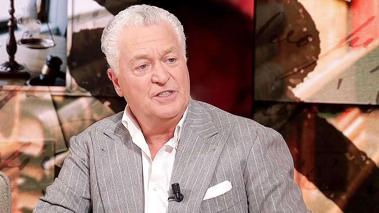 Man with silver hair in a gray pinstripe suit speaks on a tv talk show set microphone clipped to his lapel