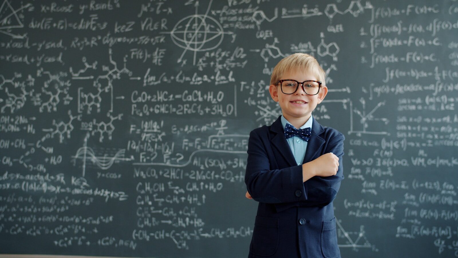 Young boy in glasses smiles in front of chalkboard equations
