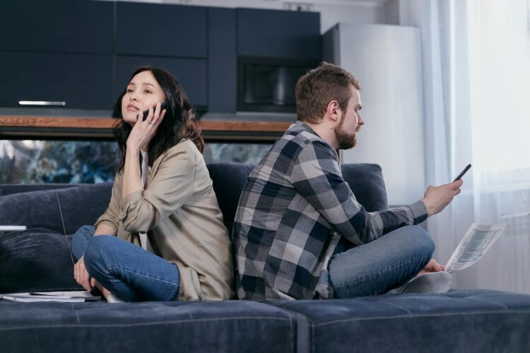 A couple sitting back to back on a sofa expressing financial tension while using phones
