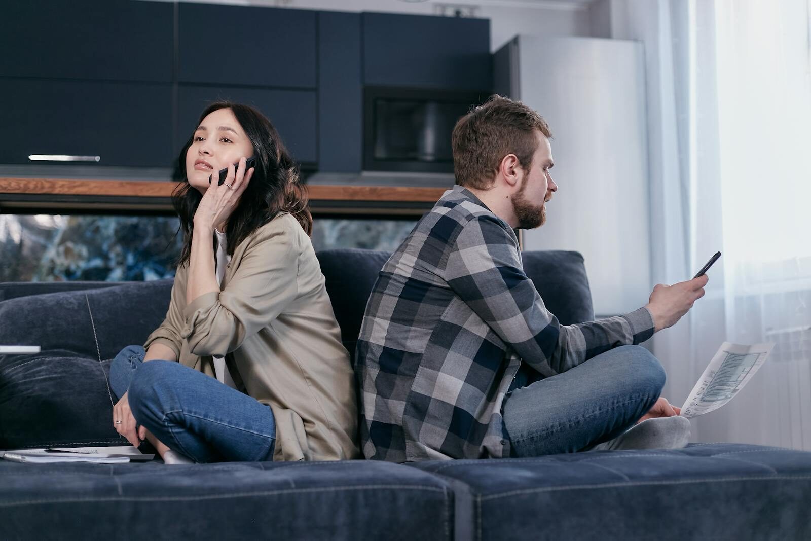 A couple sitting back to back on a sofa expressing financial tension while using phones