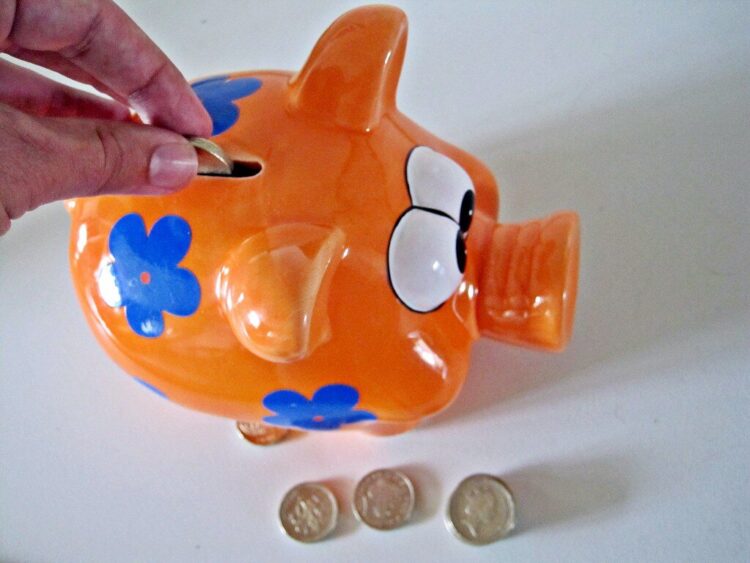 Hand inserts a coin into an orange piggy bank decorated with blue flowers with several coins on the white surface nearby