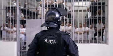 Police officer in riot gear facing prisoners behind barred doors in a detention area.