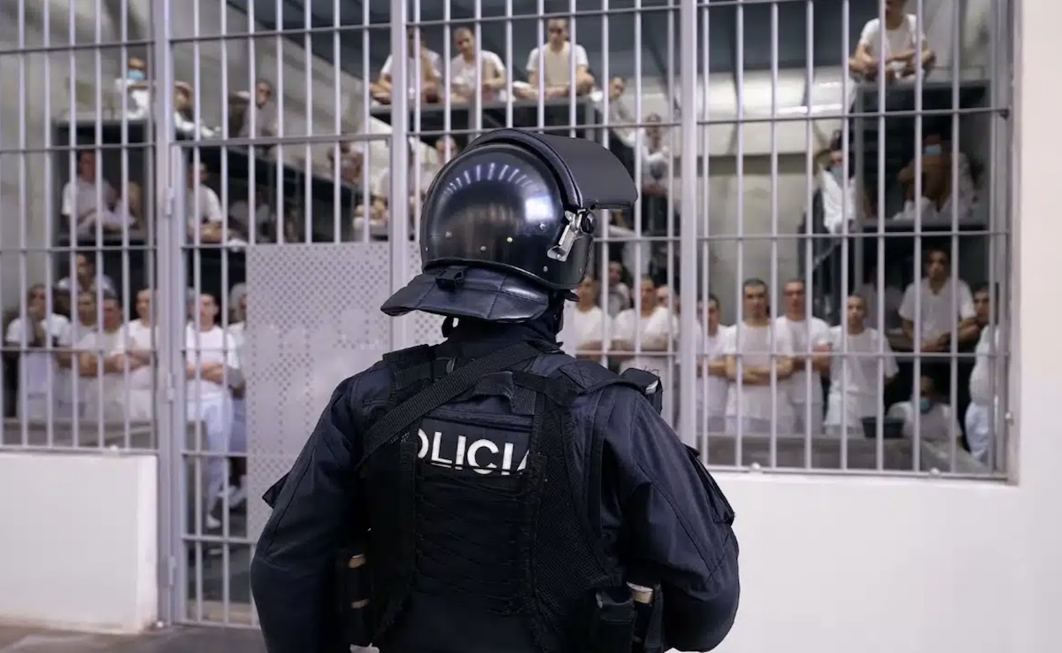 Police officer in riot gear facing prisoners behind barred doors in a detention area
