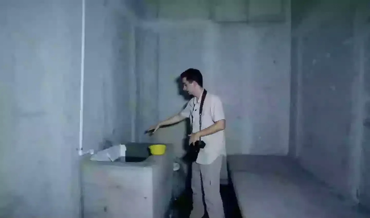 Man with a camera hanging from his neck points at objects on a concrete work surface in a bare dim room a yellow bucket sits nearby