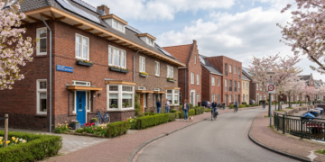 Row of brick houses along a canal street with blooming cherry trees, pedestrians and cyclists nearby.