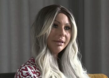 Woman with long blonde hair wearing a red and white patterned top seated indoors beside a yellow chair
