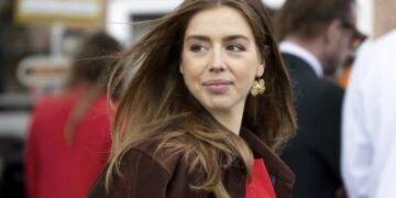 Woman with long brown hair wearing a brown coat and red blouse, looking to her left in a busy outdoor setting.