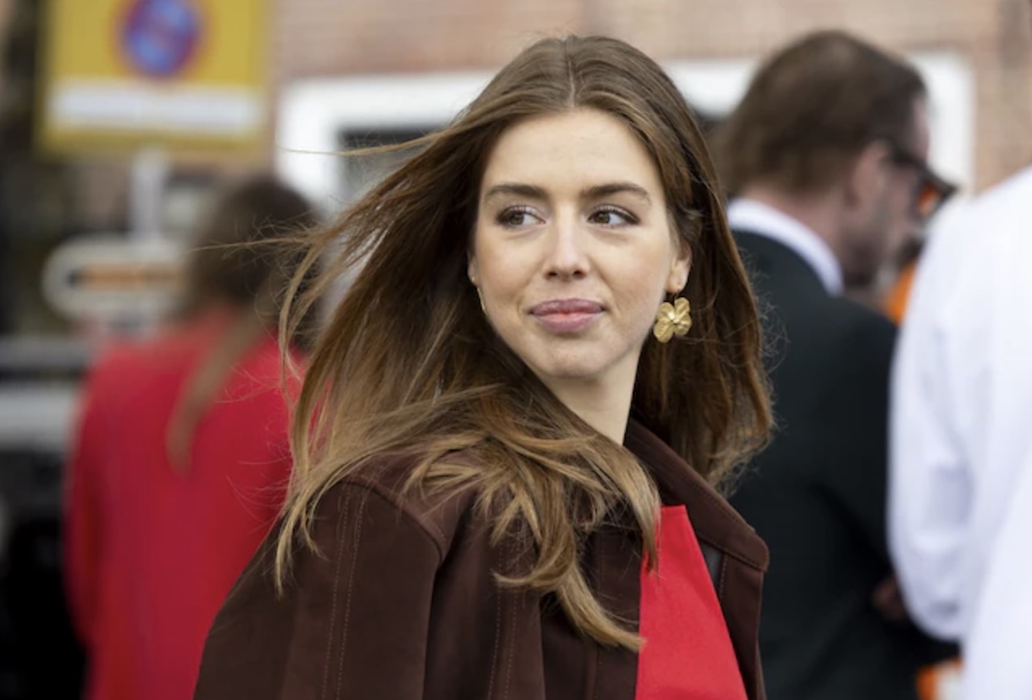 Woman with long brown hair wearing a brown coat and red blouse looking to her left in a busy outdoor setting