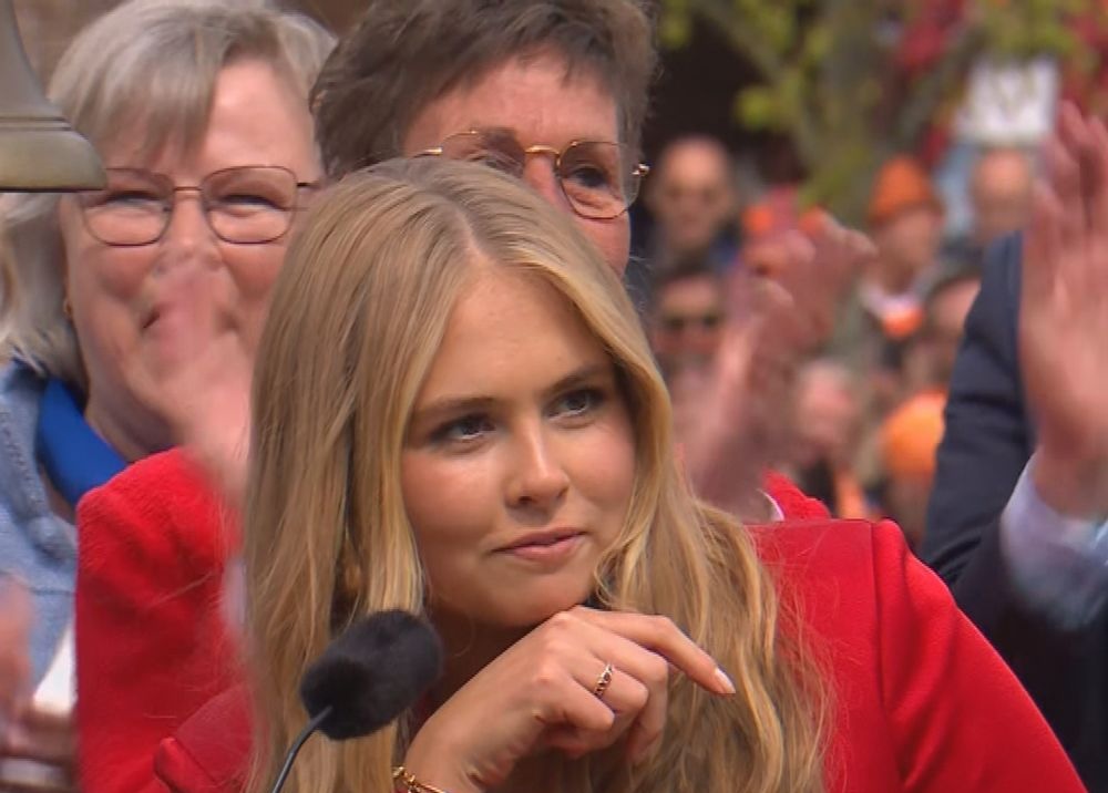 Woman in a red blazer at a microphone during a public appearance with a cheering crowd in the background