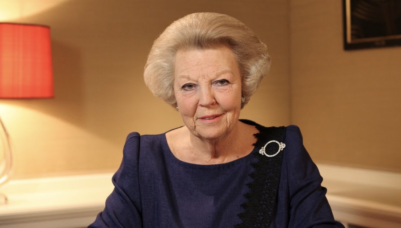 Older woman with white hair in a navy dress seated indoors warm lamp behind her