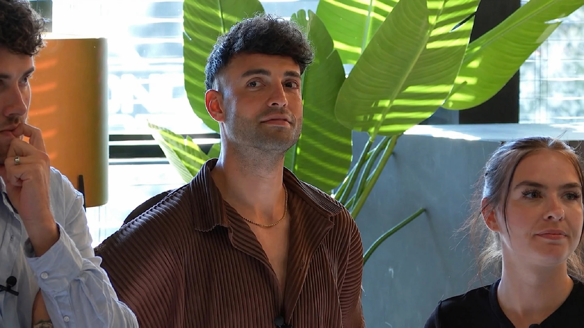 Three adults indoors beside a large tropical plant center man in a brown striped shirt looks toward the camera others partially visible on each side