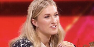 Blonde woman with a braided headband, smiling, seated against a bright red background."