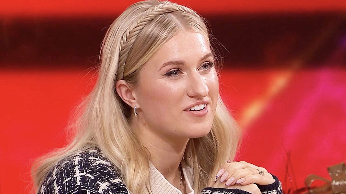 Blonde woman with a braided headband smiling seated against a bright red background 
