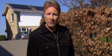 Man in a black jacket stands in a driveway beside a tall brown hedge, with a brick house, a parked car, and solar panels visible on the roof in a sunny suburban scene.