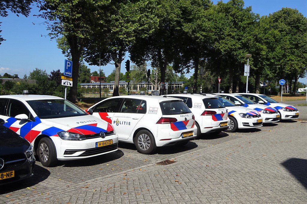 Line of dutch politie cars parked along a cobblestone street on a sunny day with blue red chevron livery and'Politie' markings.