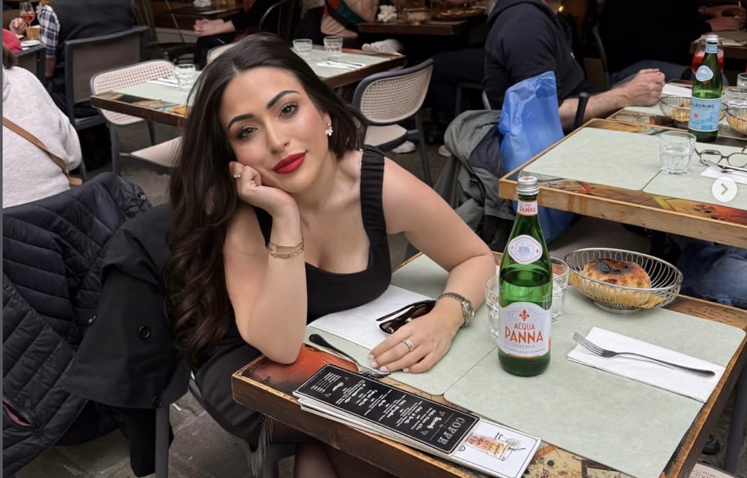 - Info Vandaag Woman with long dark hair in a black sleeveless top rests her chin on her hand at an outdoor cafe table a bottle of acqua panna and a pastry are on the table