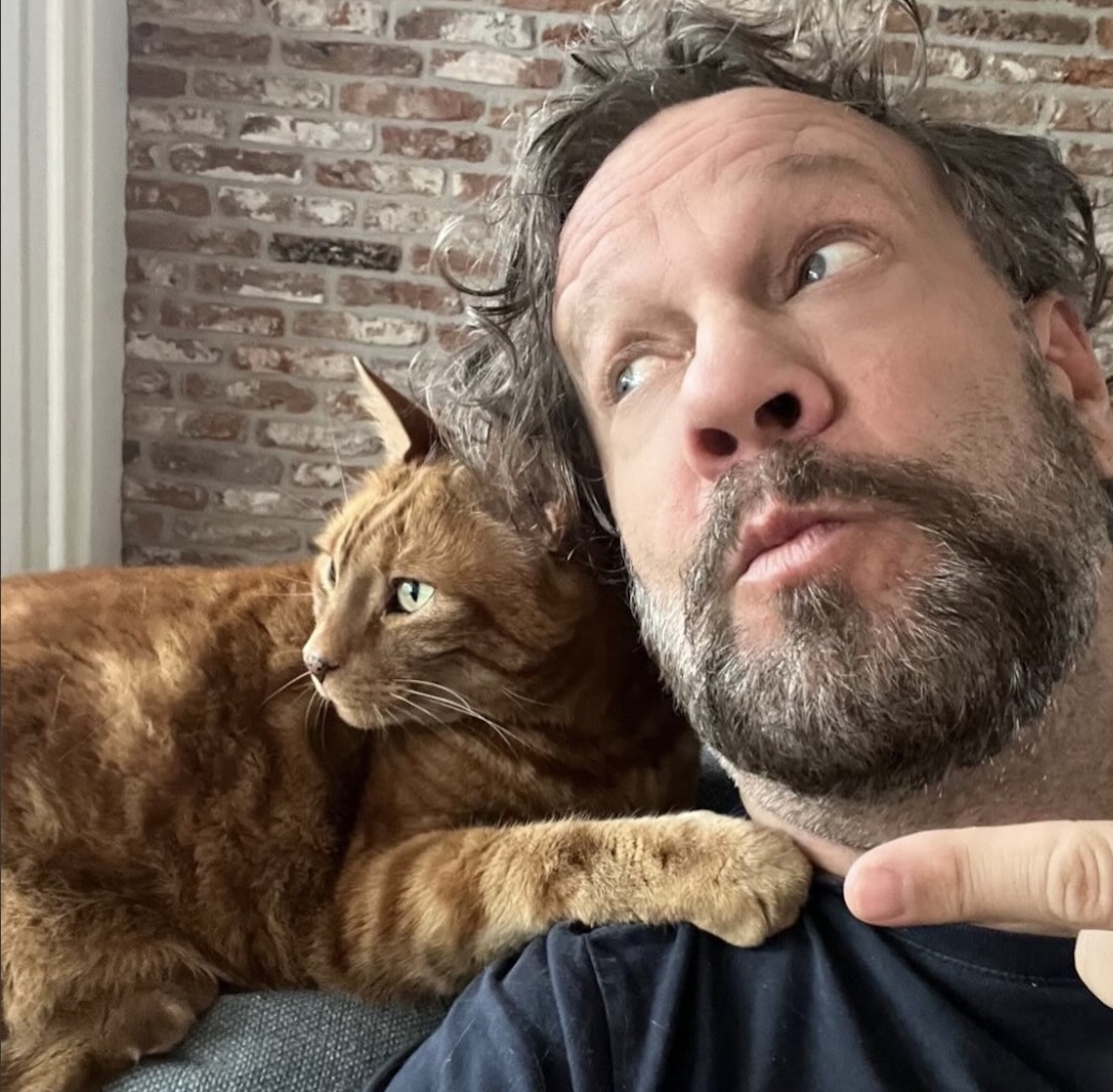 Man with a gray beard taking a selfie while an orange tabby cat rests its paw on his shoulder beside his face