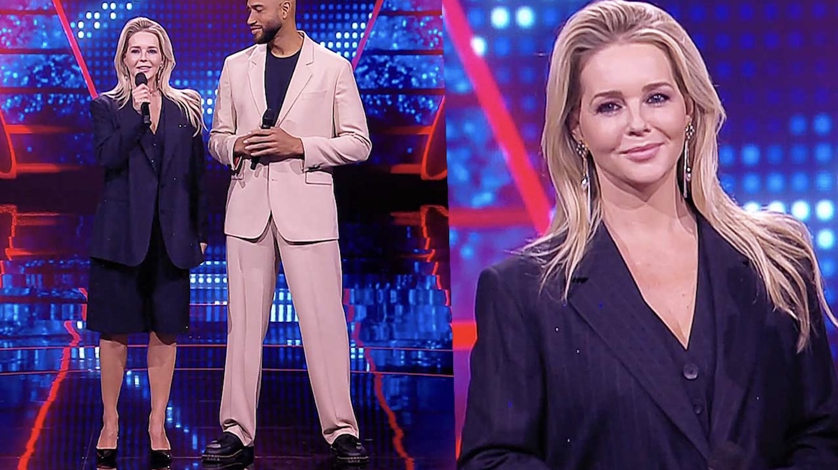 Two tv hosts a woman with a microphone and a man in a light beige suit stand on a bright blue red stage backdrop
