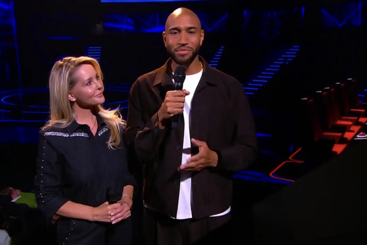 Two presenters stand on a dark stage holding handheld microphones the woman looking at the man as blue lights glow in the background