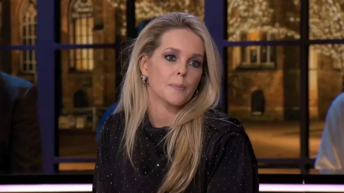Blonde woman with long hair wearing a dotted black top sits in a tv studio during a discussion looking slightly to the side