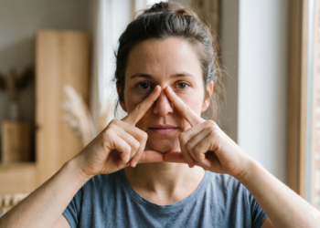 Woman in a blue shirt forms a square frame around her eyes with her fingers in front of her face by a window
