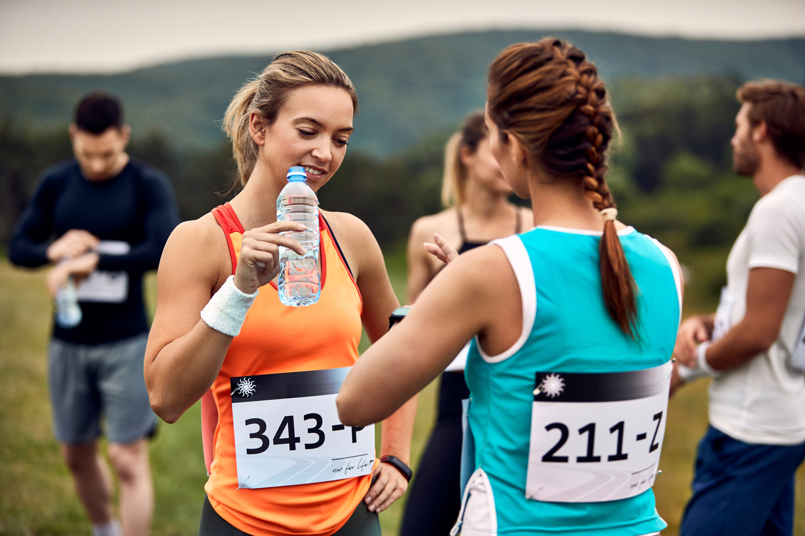 Two female runners share a water break during a race one in an orange top drinking from a bottle while the other in teal speaks nearby with race bibs visible on their waists and hills in the background