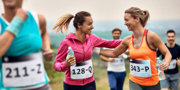 Two smiling female runners share a high-five during a race, each holding a water bottle.