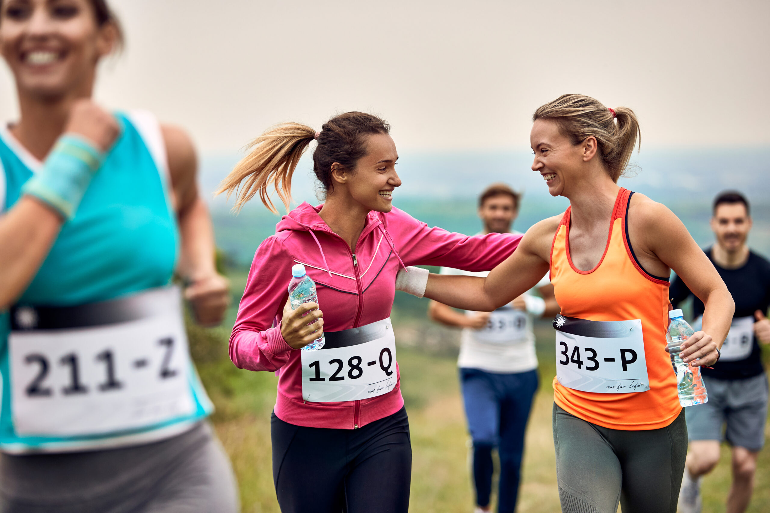 Two smiling female runners share a high five during a race each holding a water bottle