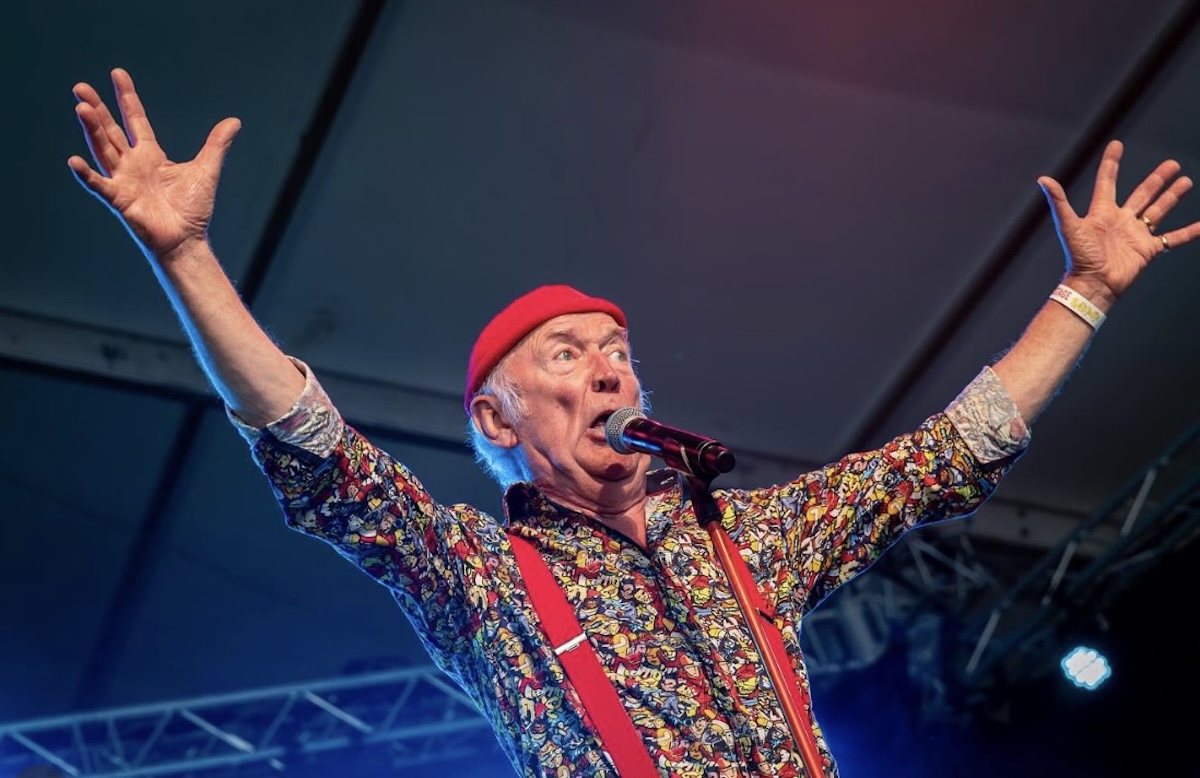 Older male singer on a stage red beret colorful shirt and red suspenders arms raised as he sings into a microphone