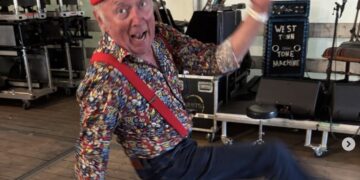 Older man in a colorful patterned shirt and red hat dancing on a stage, smiling with one arm raised.