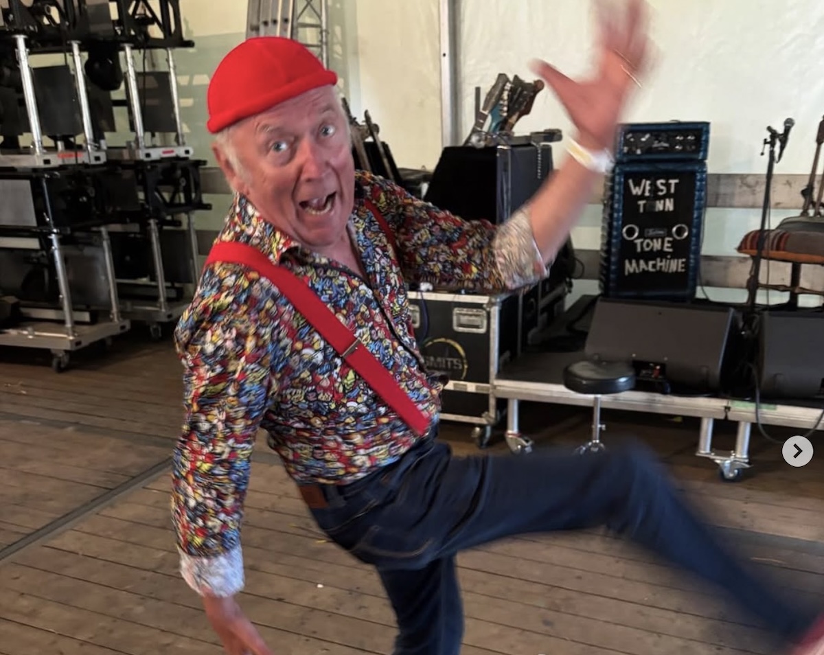 Older man in a colorful patterned shirt and red hat dancing on a stage smiling with one arm raised